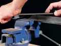 Händer filar på en metallprofil som sitter fast i ett blått skruvstycke.