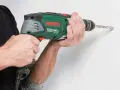 Händerna håller en grön Bosch-borrmaskin som borrar i en vit vägg.
