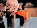 Händer använder en popnittång för att fästa en popnit. Tången är orange och svart, och popniten är silverfärgad. Det här är ett exempel på hur man arbetar med popnit dimensioner.