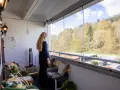 En kvinna njuter av utsikten från sin inredda lilla balkong med glaspartier. Våren syns i träden utanför.