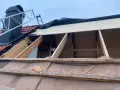 Taket är under renovering. Träbjälkar och takpannor syns, samt en stege vid skorstenen. Arbetet med att laga taket pågår.