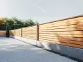 Ett modernt dubbelsidigt plank stående med horisontella träribbor och grå betongfundament. En smal cypress i kruka står vid staketet.