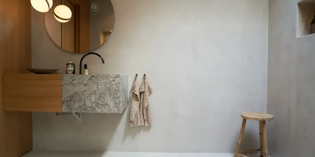 Modernt badrum med handfat i marmor och trä, rund spegel och två lampor. Tänk på microcement badrum pris för en liknande stil.