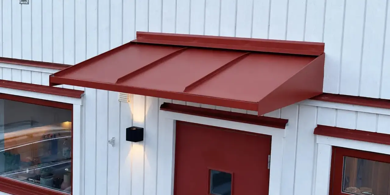 Ett rött plåttak i gammal stil skyddar en dörr och ett fönster på ett vitt trähus.