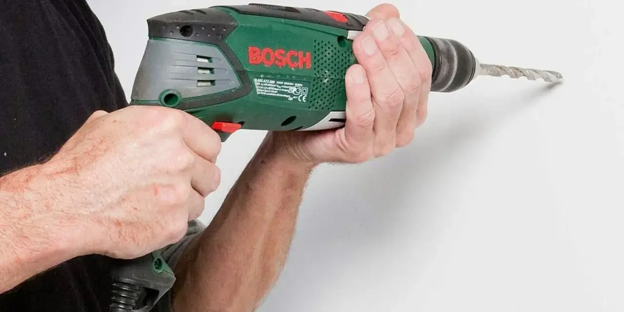 Händerna håller en grön Bosch-borrmaskin som borrar i en vit vägg.