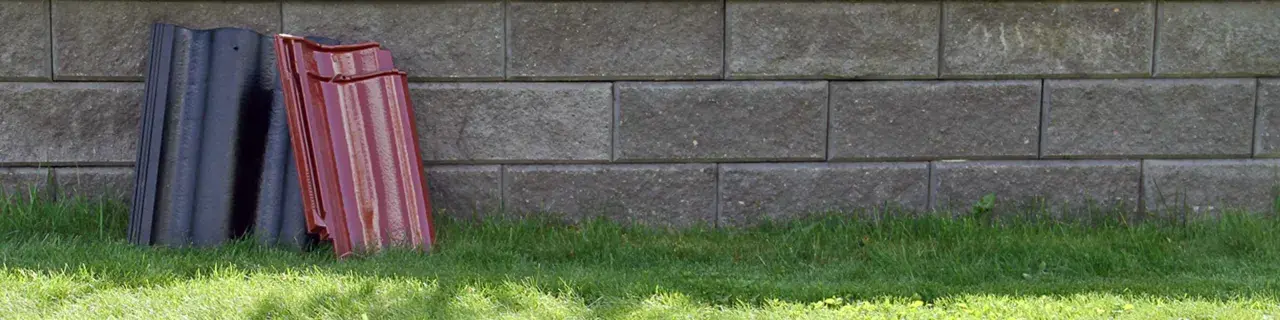 Två staplar takpannor, en svart och en röd, lutar mot en mur av grå betongblock. Undrar vad kostar takpannor?