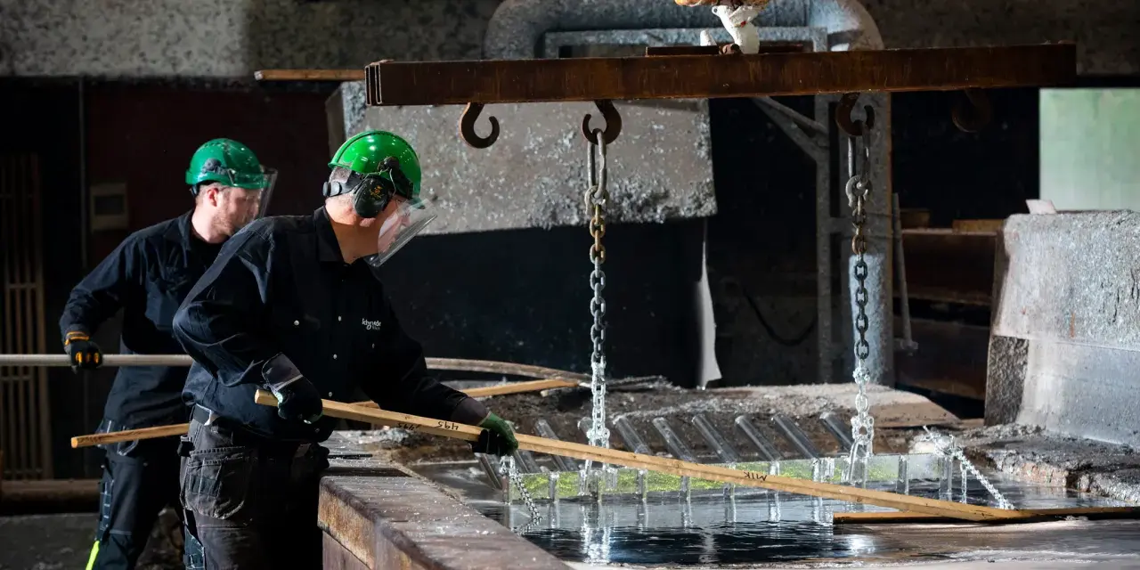 Två arbetare i skyddsutrustning hanterar metall som doppas i ett bad. Processen för varmförzinkning pris kan variera beroende på material och storlek.
