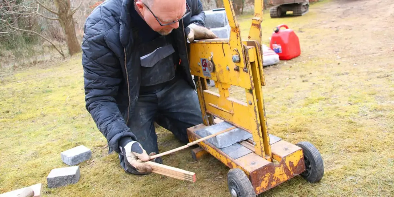 Man klipper marksten med en gul maskin. Han mäter med tumstocken för att få rätt längd.