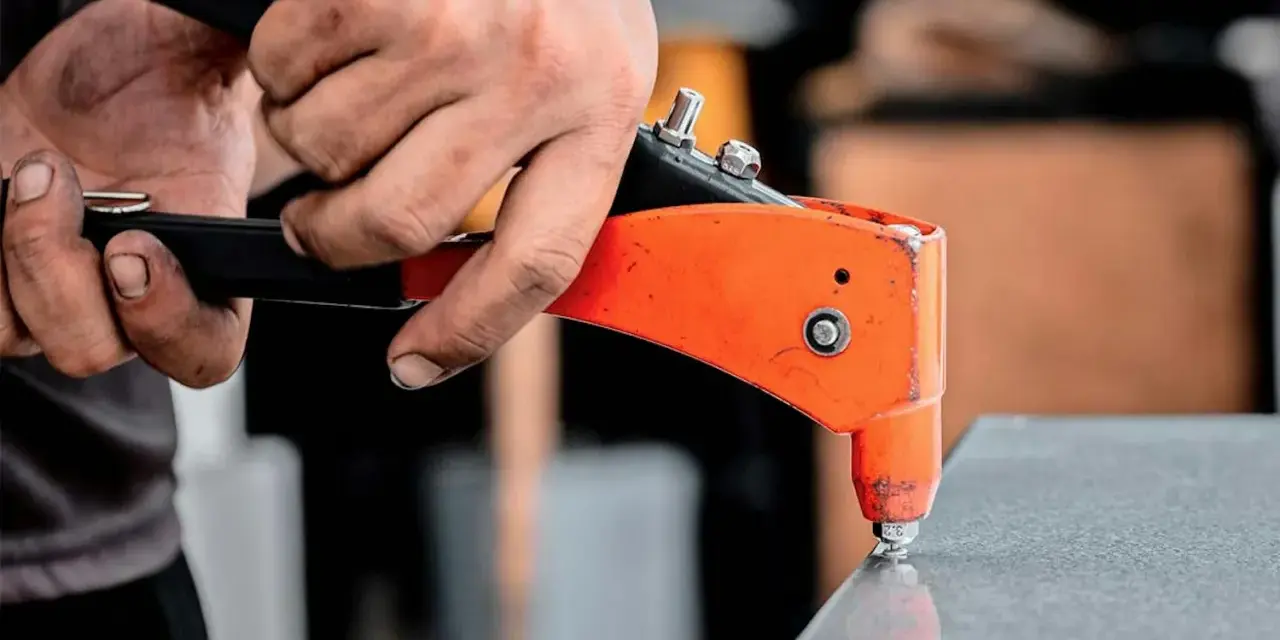 Händer använder en popnittång för att fästa en popnit. Tången är orange och svart, och popniten är silverfärgad. Det här är ett exempel på hur man arbetar med popnit dimensioner.