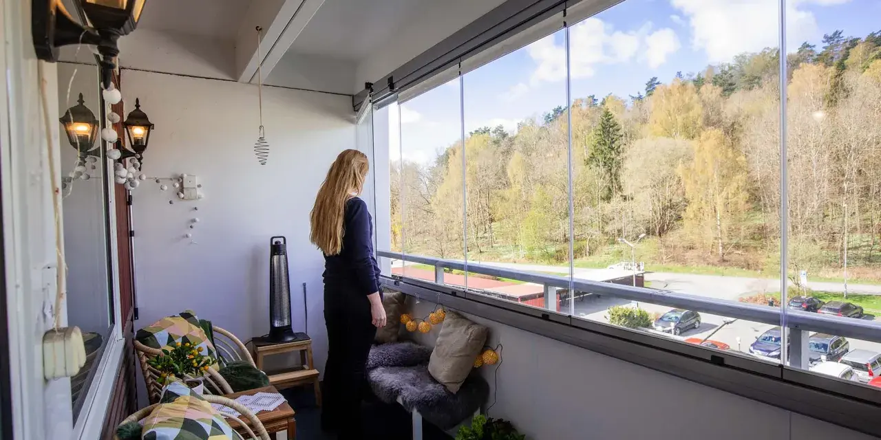 En kvinna njuter av utsikten från sin inredda lilla balkong med glaspartier. Våren syns i träden utanför.