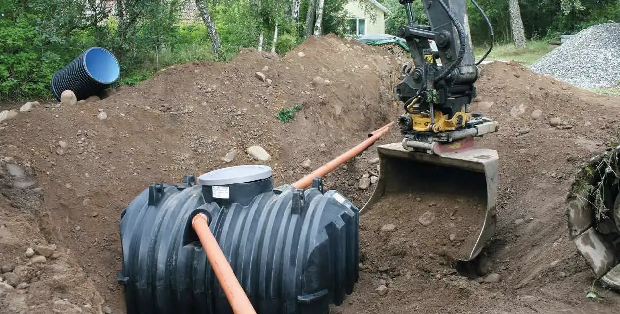 Grävmaskin installerar enskilt avlopp. En stor plasttank placeras i marken, vilket är en del av enskilt avlopp kostnad.