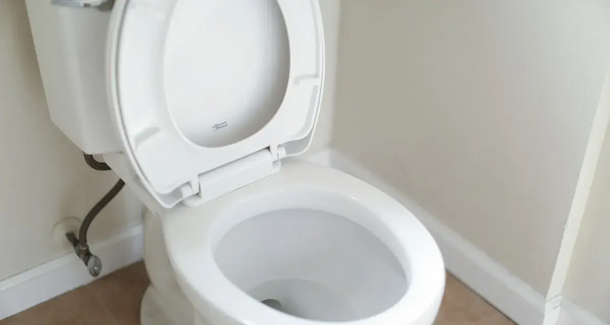 En vit toalett med öppet lock. Vattenanslutningen syns vid sidan, vilket kan tyda på ett potentiellt stopp i toaletten.
