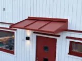 Ett rött plåttak i gammal stil skyddar en dörr och ett fönster på ett vitt trähus.
