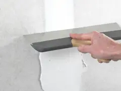 Handen spacklar en vägg med en bred spackel. Arbetet med att kapa gipsskiva kan nu fortsätta.