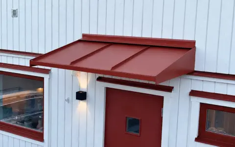 Ett rött plåttak i gammal stil skyddar en dörr och ett fönster på ett vitt trähus.