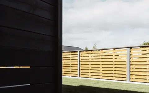 En modern, horisontell trästaket som ger en känsla av att bygga billigt staket. Grön gräsmatta och en mörk vägg i förgrunden.
