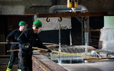 Två arbetare i skyddsutrustning hanterar metall som doppas i ett bad. Processen för varmförzinkning pris kan variera beroende på material och storlek.