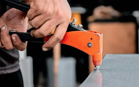 Händer använder en popnittång för att fästa en popnit. Tången är orange och svart, och popniten är silverfärgad. Det här är ett exempel på hur man arbetar med popnit dimensioner.