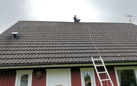 En person sitter på ett tegeltak med en stege bredvid. Taket är täckt av mossa, vilket gör det svårt att avgöra hur många tegelpannor per kvadratmeter som används.