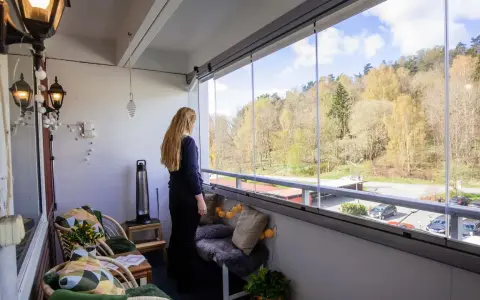 En kvinna njuter av utsikten från sin inredda lilla balkong med glaspartier. Våren syns i träden utanför.