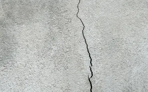 En spricka löper genom en grov, grå putsad fasad. Dags att putsa fasaden för att laga skadan.