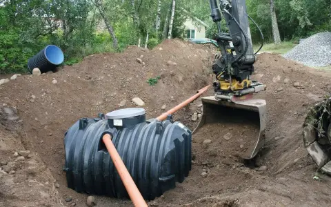 Grävmaskin installerar enskilt avlopp. En stor plasttank placeras i marken, vilket är en del av enskilt avlopp kostnad.