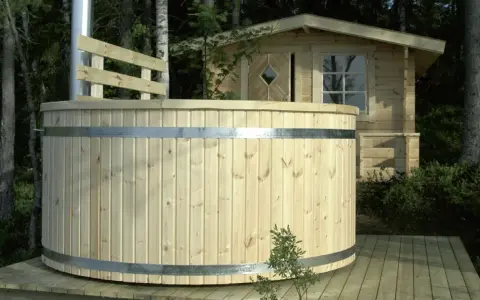 En nybyggd badtunna i trä står på en altan i skogen. Bredvid syns en liten timrad stuga.