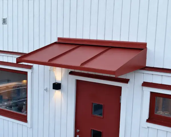Ett rött plåttak i gammal stil skyddar en dörr och ett fönster på ett vitt trähus.