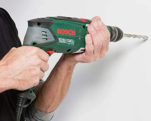 Händerna håller en grön Bosch-borrmaskin som borrar i en vit vägg.