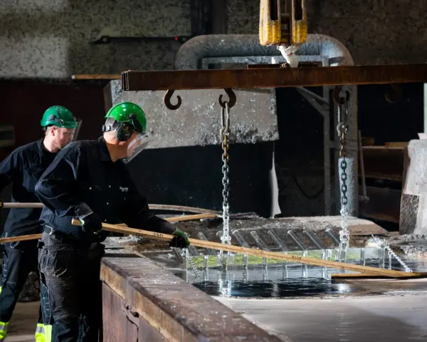 Två arbetare i skyddsutrustning hanterar metall som doppas i ett bad. Processen för varmförzinkning pris kan variera beroende på material och storlek.