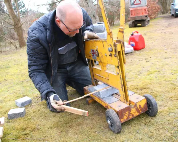 Man klipper marksten med en gul maskin. Han mäter med tumstocken för att få rätt längd.