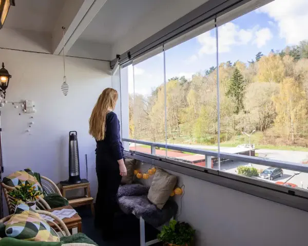 En kvinna njuter av utsikten från sin inredda lilla balkong med glaspartier. Våren syns i träden utanför.