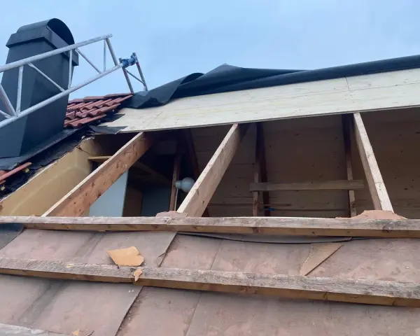 Taket är under renovering. Träbjälkar och takpannor syns, samt en stege vid skorstenen. Arbetet med att laga taket pågår.
