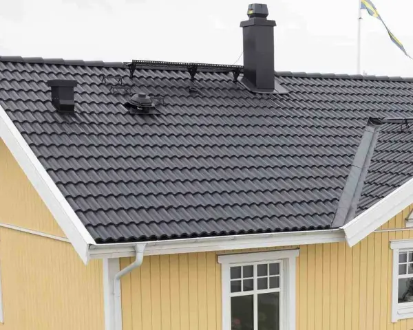 Taket har mörka takpannor, en svart skorsten och en svensk flagga. Vindskiveplåt montering syns längs takkanten.