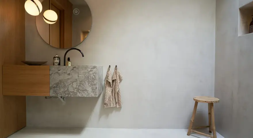Modernt badrum med handfat i marmor och trä, rund spegel och två lampor. Tänk på microcement badrum pris för en liknande stil.