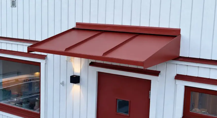 Ett rött plåttak i gammal stil skyddar en dörr och ett fönster på ett vitt trähus.