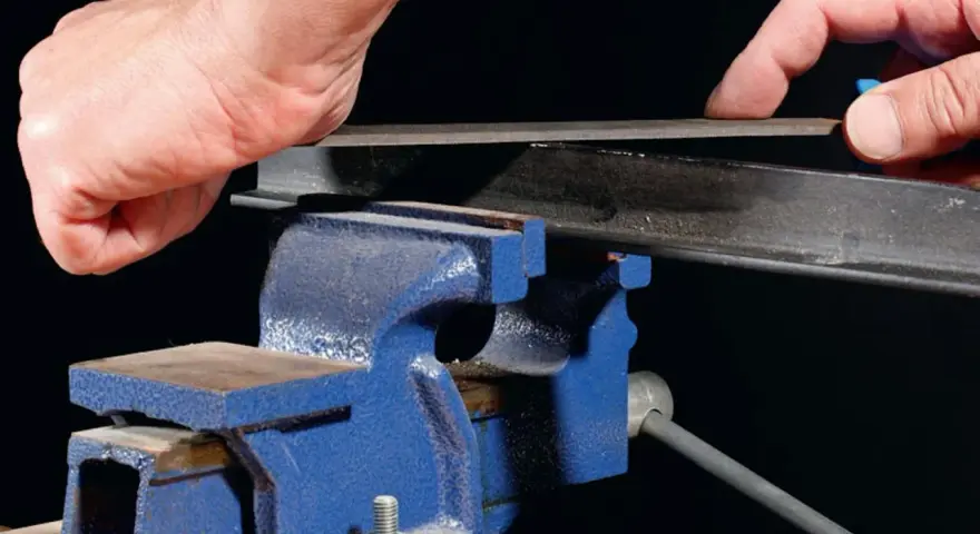 Händer filar på en metallprofil som sitter fast i ett blått skruvstycke.