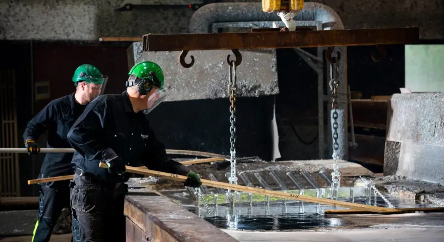 Två arbetare i skyddsutrustning hanterar metall som doppas i ett bad. Processen för varmförzinkning pris kan variera beroende på material och storlek.