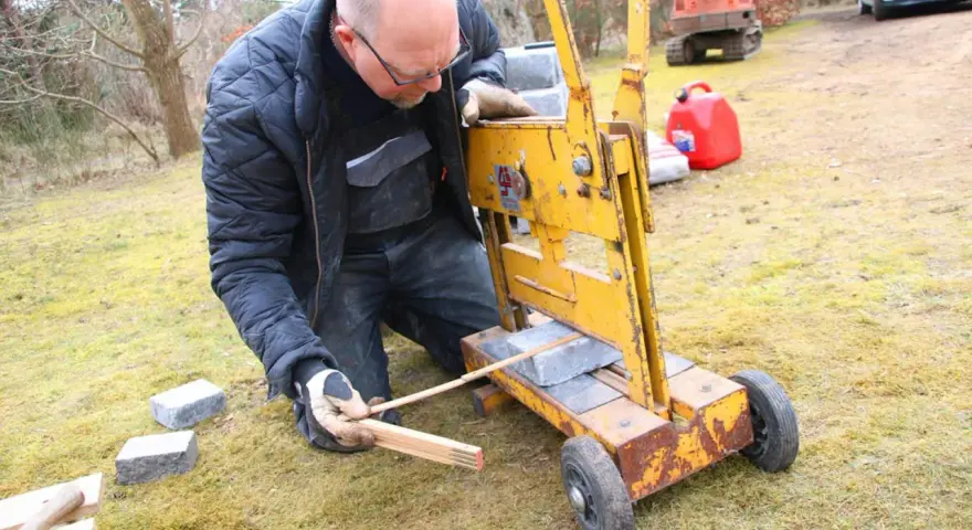 Man klipper marksten med en gul maskin. Han mäter med tumstocken för att få rätt längd.