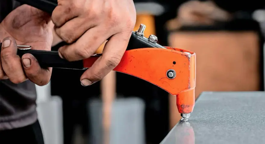 Händer använder en popnittång för att fästa en popnit. Tången är orange och svart, och popniten är silverfärgad. Det här är ett exempel på hur man arbetar med popnit dimensioner.