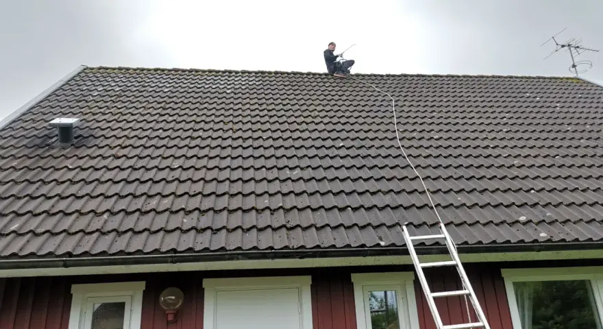 En person sitter på ett tegeltak med en stege bredvid. Taket är täckt av mossa, vilket gör det svårt att avgöra hur många tegelpannor per kvadratmeter som används.