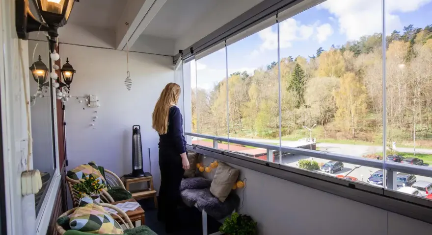 En kvinna njuter av utsikten från sin inredda lilla balkong med glaspartier. Våren syns i träden utanför.