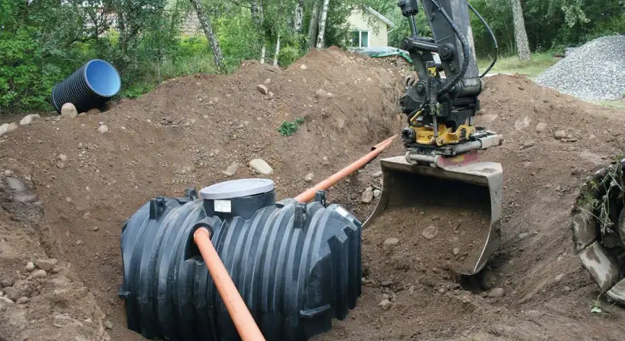 Grävmaskin installerar enskilt avlopp. En stor plasttank placeras i marken, vilket är en del av enskilt avlopp kostnad.