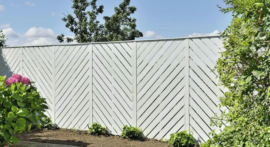 Vitt staket med diagonalt mönster ger staket inspiration. Vackert mot blå himmel och grönska.