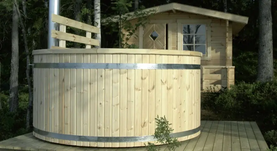 En nybyggd badtunna i trä står på en altan i skogen. Bredvid syns en liten timrad stuga.