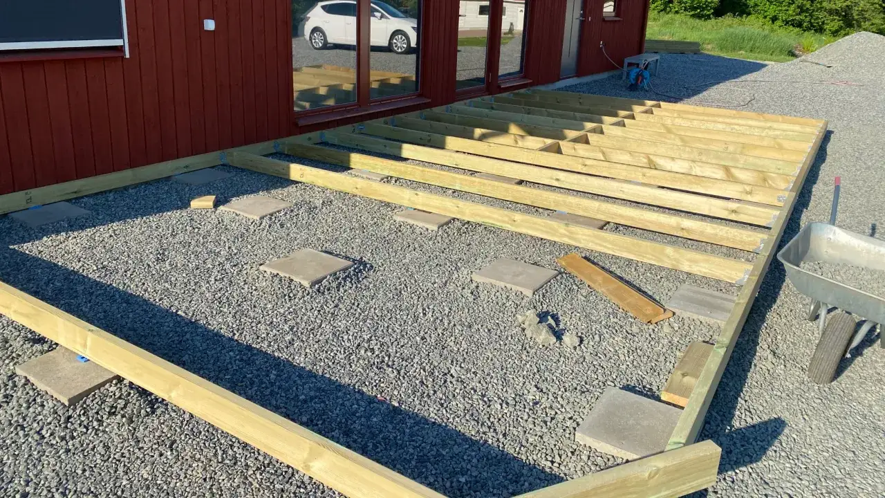 Grundarbete för att bygga altan på mark. Träreglar ligger redo att monteras på grusunderlag.