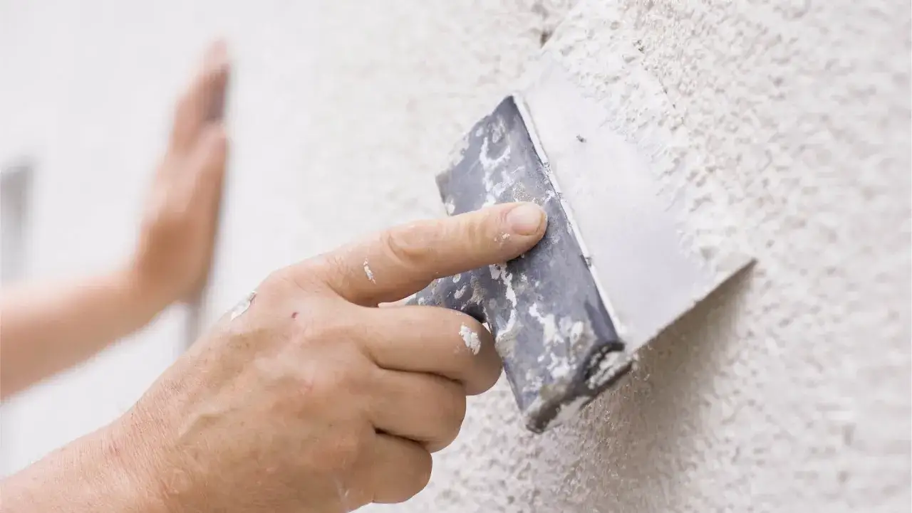 En hand med en spackel drar över en texturerad vägg, en del av en fasadrenovering puts.
