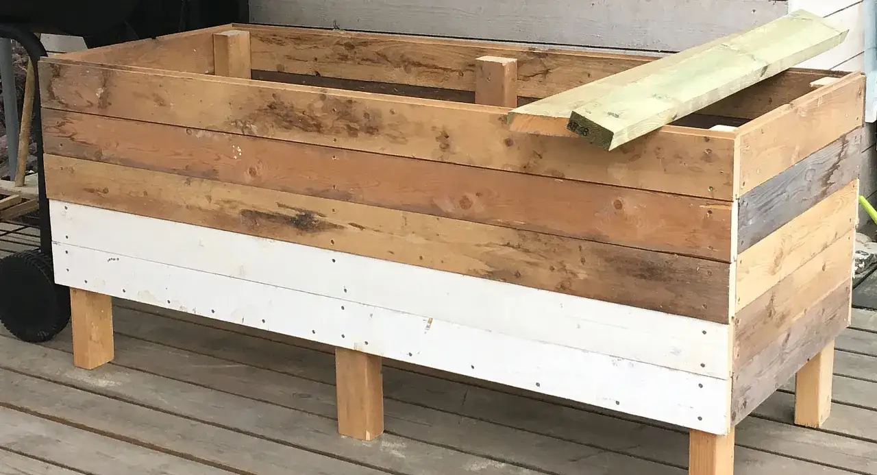 Bygga odlingslåda med botten. En rektangulär träkonstruktion med flera lager av träplankor, varav en är vitmålad. En ny träplanka ligger ovanpå.