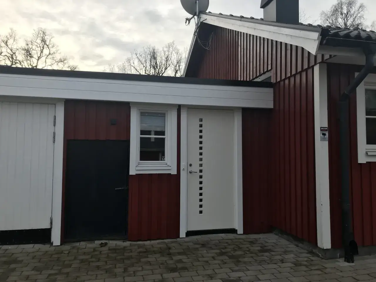 Rött hus med vita vindskivor och takrännor. En vit dörr med fyrkantiga fönster och en svart dörr syns.