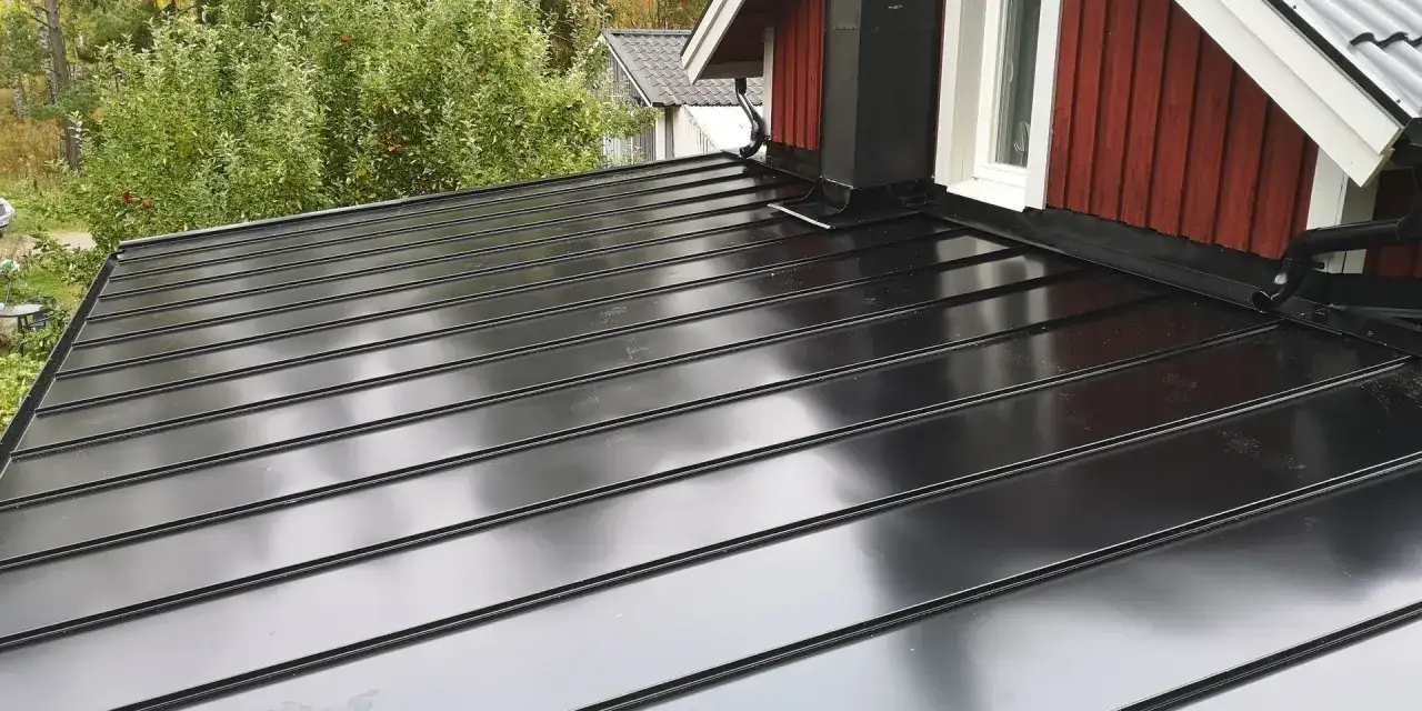 Ett glänsande, svart plåttak med synliga falsar. En skorsten och stuprör i svart plåt syns vid husknuten. Olika typer av plåttak kan ge ett modernt utseende.